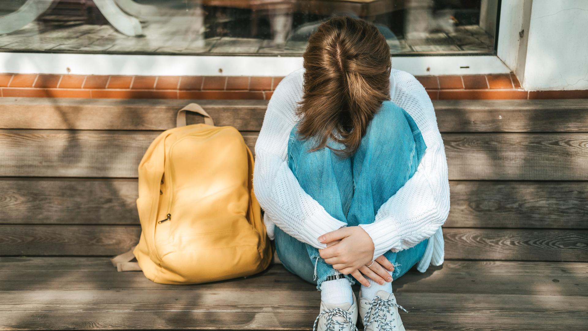 Eine Schülerin kauert neben ihrem Rucksack auf einer Bank und stützt den Kopf auf ihre Knie.