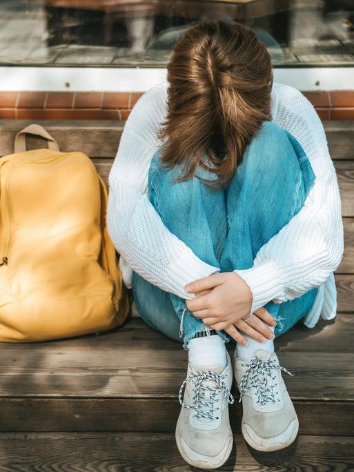 Eine Schülerin kauert neben ihrem Rucksack auf einer Bank und stützt den Kopf auf ihre Knie.