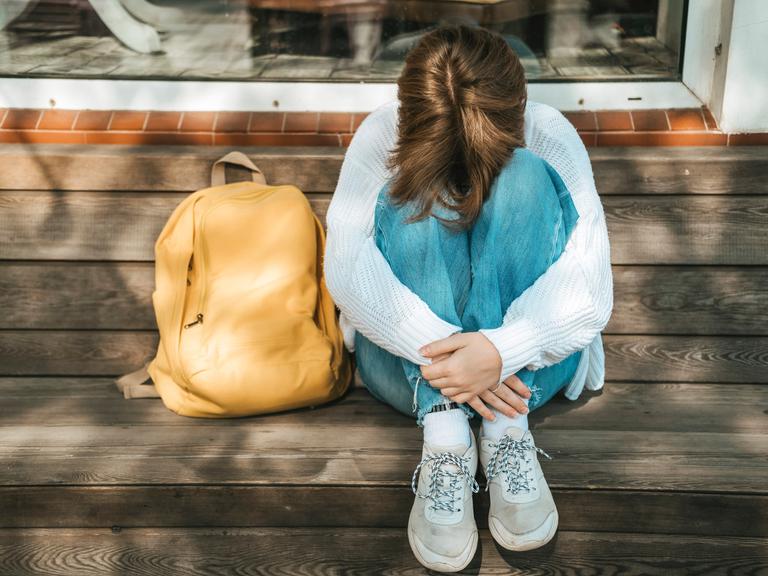 Eine Schülerin kauert neben ihrem Rucksack auf einer Bank und stützt den Kopf auf ihre Knie.