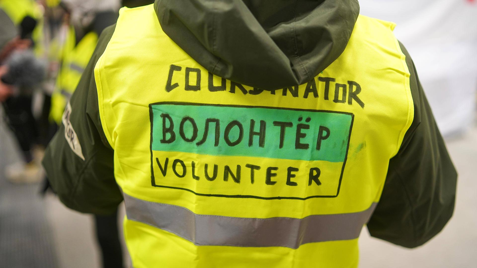 Rückenansicht einer Person in einer neon gelben Weste, mit der Aufschrift "Volunteer", in englischer und auch in russischer Sprache.