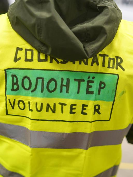 Rückenansicht einer Person in einer neon gelben Weste, mit der Aufschrift "Volunteer", in englischer und auch in russischer Sprache.