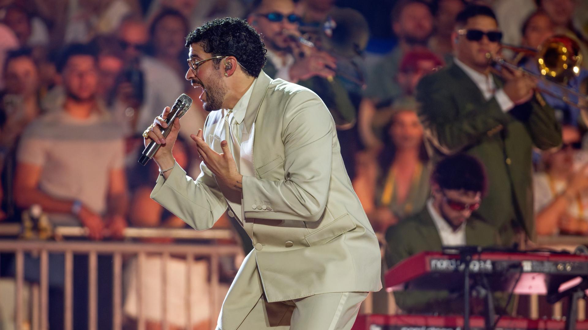 Bad Bunny steht auf der Bühne und singt in ein Mikrofon. Er trägt einen beigefarbenen Anzug. Im Hintergrund stehen Musiker und Fans. Bad Bunny steht auf der Bühne und singt in ein Mikrofon. Er trägt einen beigefarbenen Anzug. Im Hintergrund stehen Musiker und Fans.