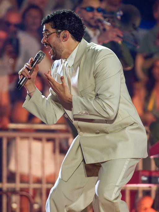 Bad Bunny steht auf der Bühne und singt in ein Mikrofon. Er trägt einen beigefarbenen Anzug. Im Hintergrund stehen Musiker und Fans.