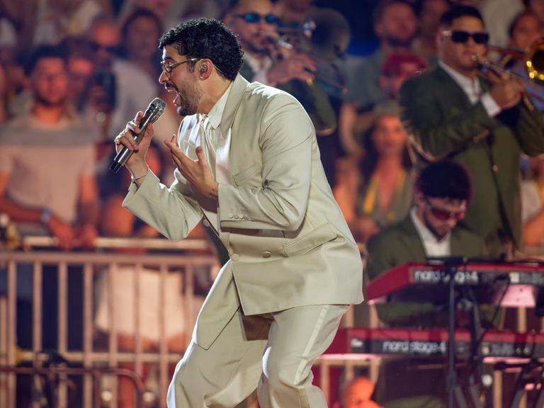 Bad Bunny steht auf der Bühne und singt in ein Mikrofon. Er trägt einen beigefarbenen Anzug. Im Hintergrund stehen Musiker und Fans.