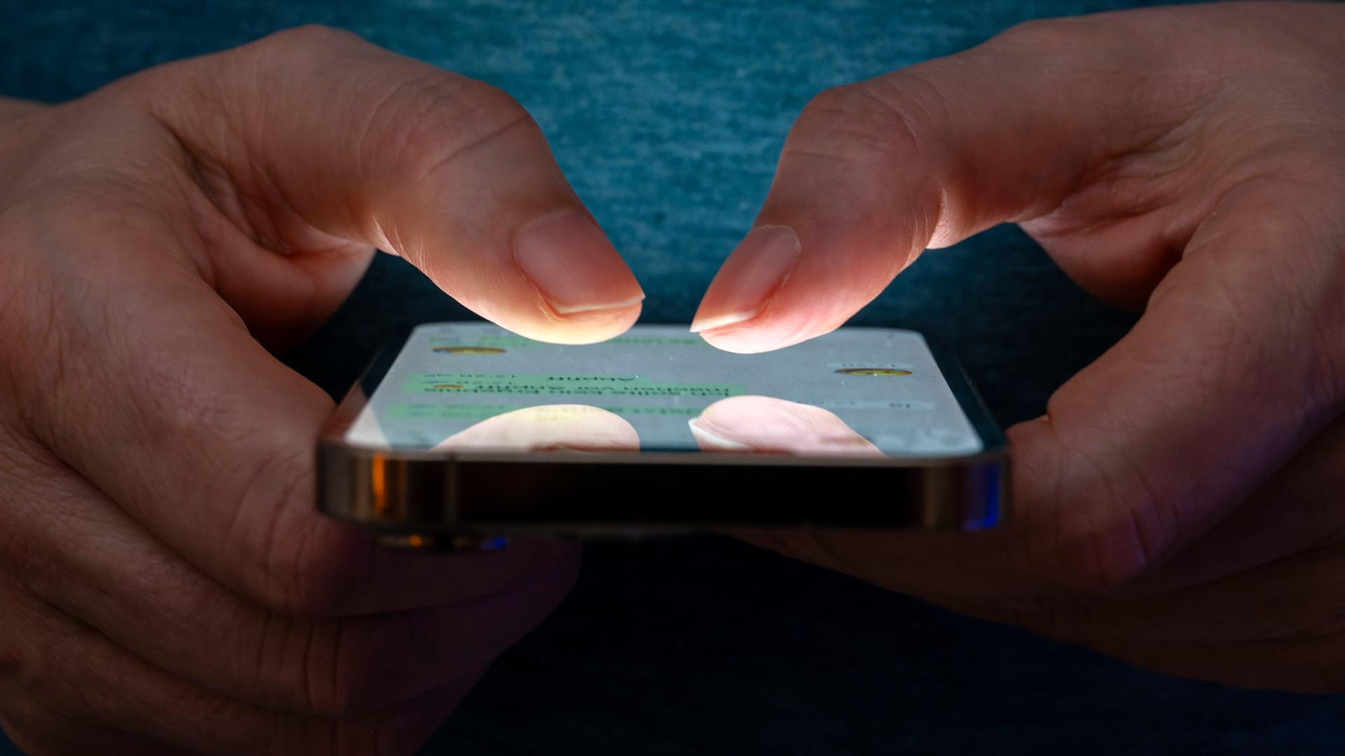Eine Frau tippt auf einem Smartphone-Screen. Das Bild zeigt eine Person, die in einer schlecht beleuchteten Umgebung ein Smartphone benutzt. Die Beleuchtung hebt die Finger des Benutzers und den Bildschirm des Smartphones hervor. Eine Frau tippt auf einem Smartphone-Screen. Das Bild zeigt eine Person, die in einer schlecht beleuchteten Umgebung ein Smartphone benutzt. Die Beleuchtung hebt die Finger des Benutzers und den Bildschirm des Smartphones hervor.