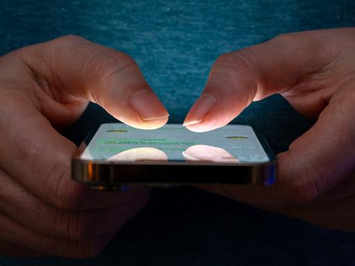 Eine Frau tippt auf einem Smartphone-Screen. Das Bild zeigt eine Person, die in einer schlecht beleuchteten Umgebung ein Smartphone benutzt. Die Beleuchtung hebt die Finger des Benutzers und den Bildschirm des Smartphones hervor.