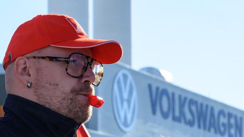 Ein Beschäftigter von Volkswagen hat eine rote Trillerpfeife im Mund und eine rote Baseball-Cappy auf dem Kopf und steht vor dem Volkswagen-Schriftzug