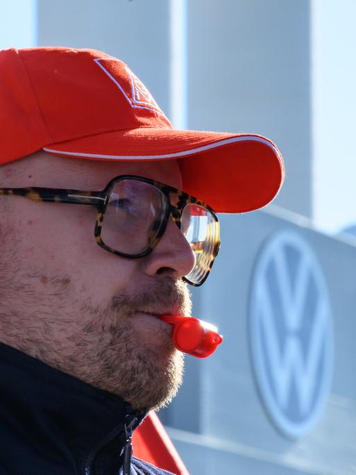 Ein Beschäftigter von Volkswagen hat eine rote Trillerpfeife im Mund und eine rote Baseball-Cappy auf dem Kopf und steht vor dem Volkswagen-Schriftzug
