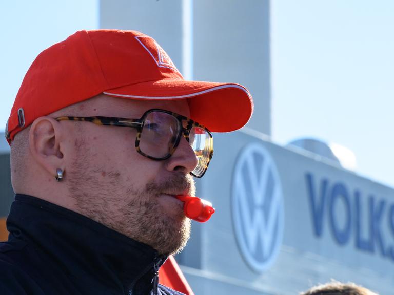 Ein Beschäftigter von Volkswagen hat eine rote Trillerpfeife im Mund und eine rote Baseball-Cappy auf dem Kopf und steht vor dem Volkswagen-Schriftzug