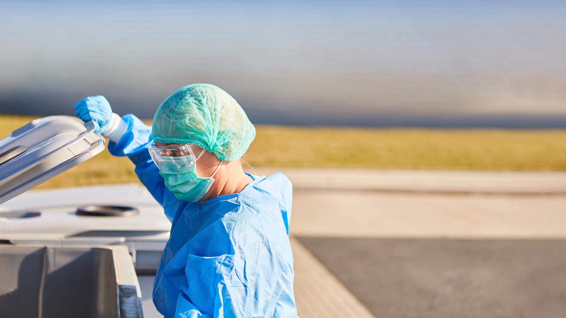 Die Putzkraft einer Klinik in Schutzkleidung öffnet eine Mülltonne.