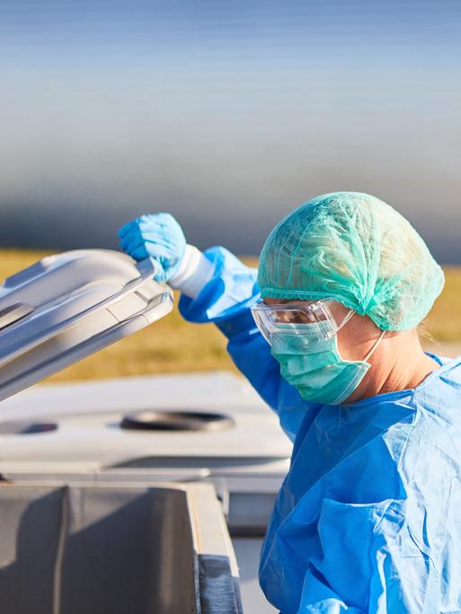 Die Putzkraft einer Klinik in Schutzkleidung öffnet eine Mülltonne.