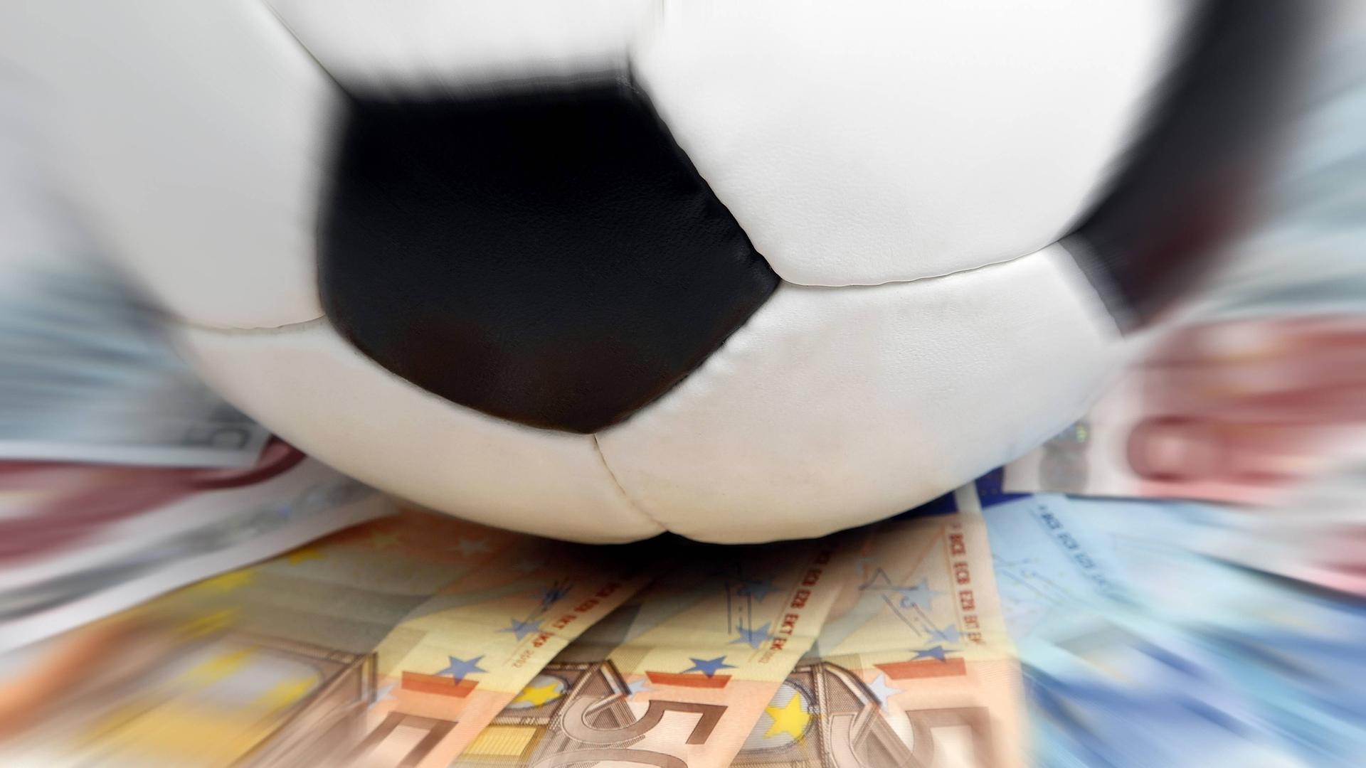 Ein Fußball rollt über Geldscheine, die auf einem Tisch liegen. 