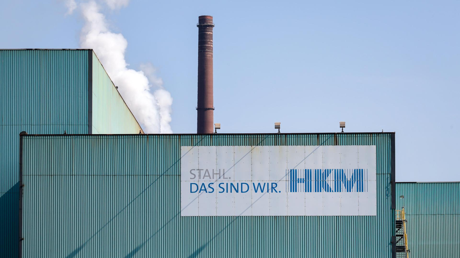 Fassade des Hüttenwerks Krupp Mannesmann in Duisburg. Vorne ein Schild mit "HKM" und dem Slogan "Stahl, das sind wir". Im Hintergrund ein brauner Schornstein.