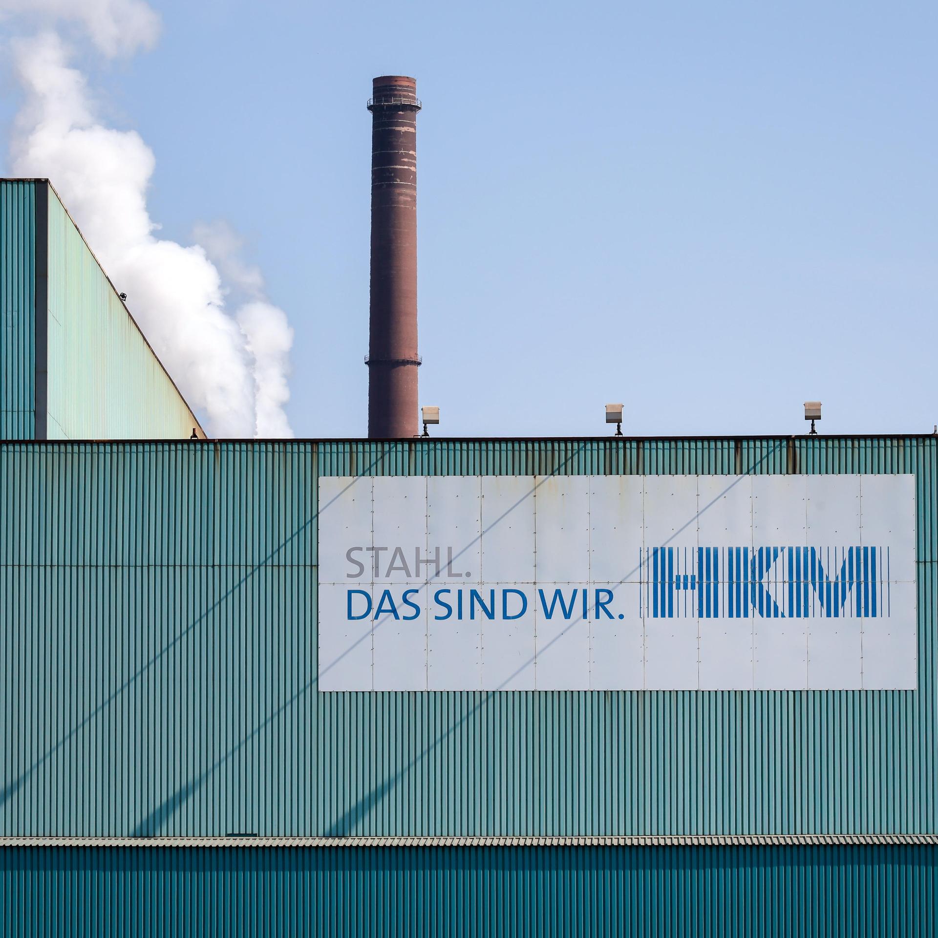 Fassade des Hüttenwerks Krupp Mannesmann in Duisburg. Vorne ein Schild mit "HKM" und dem Slogan "Stahl, das sind wir". Im Hintergrund ein brauner Schornstein.