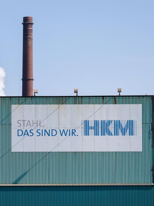 Fassade des Hüttenwerks Krupp Mannesmann in Duisburg. Vorne ein Schild mit "HKM" und dem Slogan "Stahl, das sind wir". Im Hintergrund ein brauner Schornstein.