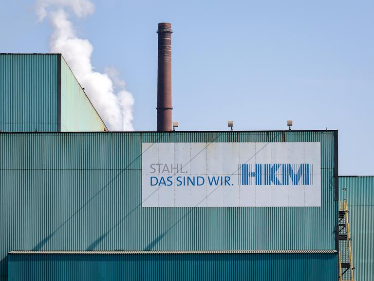 Fassade des Hüttenwerks Krupp Mannesmann in Duisburg. Vorne ein Schild mit "HKM" und dem Slogan "Stahl, das sind wir". Im Hintergrund ein brauner Schornstein.