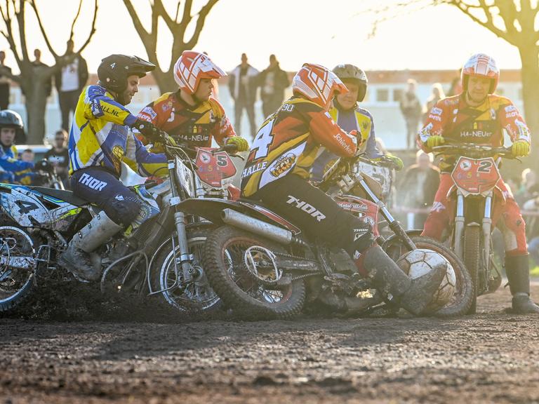 Mehrere Feldspieler auf Motorrädern wetteifern bei einer Motoball-Partie in Halle um den übergroßen Ball. Im Hintergrund sind Zuschauer zu sehen.