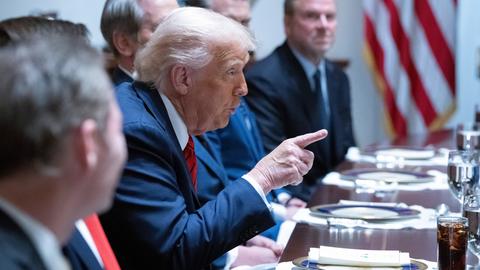 Ein Mann sitzt zwischen anderen Menschen und zeigt mit dem Zeigefinger der rechten Hand vor sich. Es ist US-Präsident Donald Trump. Er trägt einen blauen Anzug.