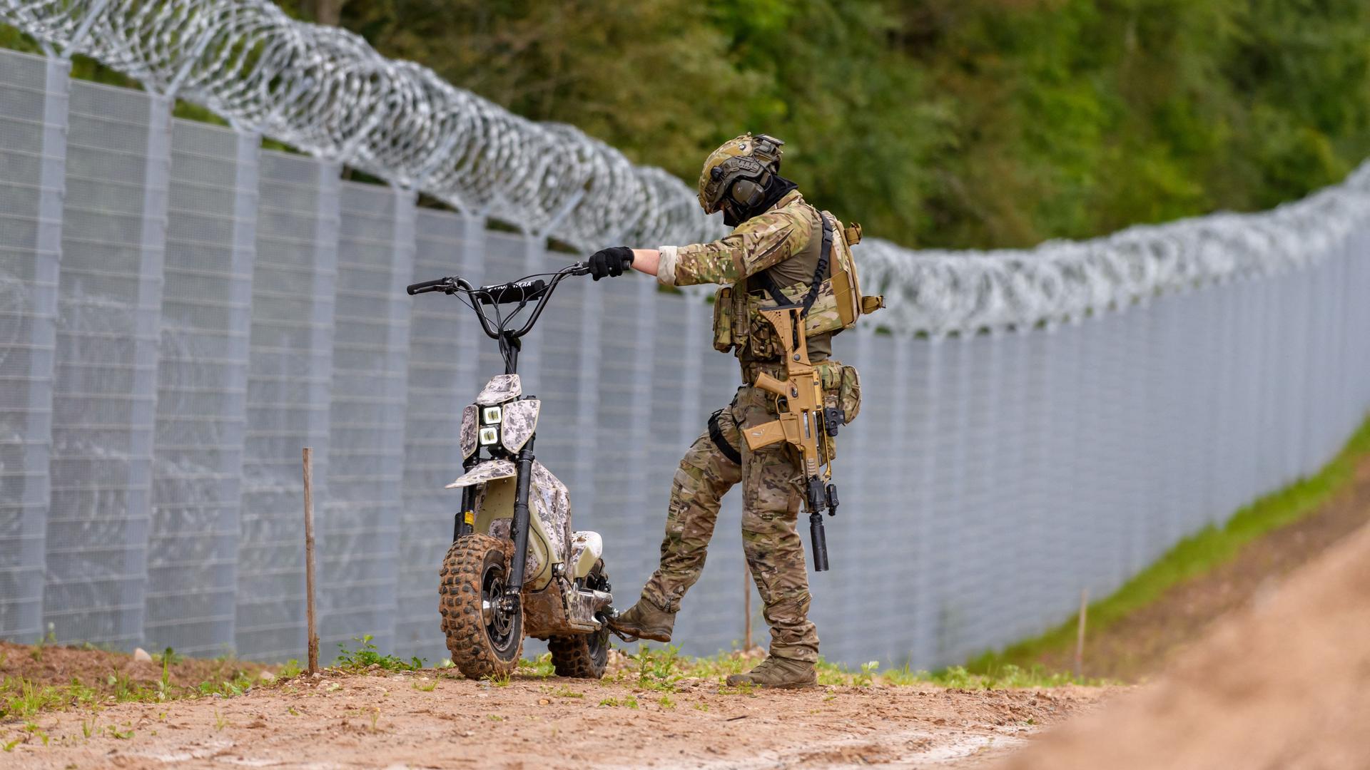 Ein Soldat der lettischen Grenzpolizei steht mit einem kleinen Motorrad am Zaun an der Grenze zu Belarus.