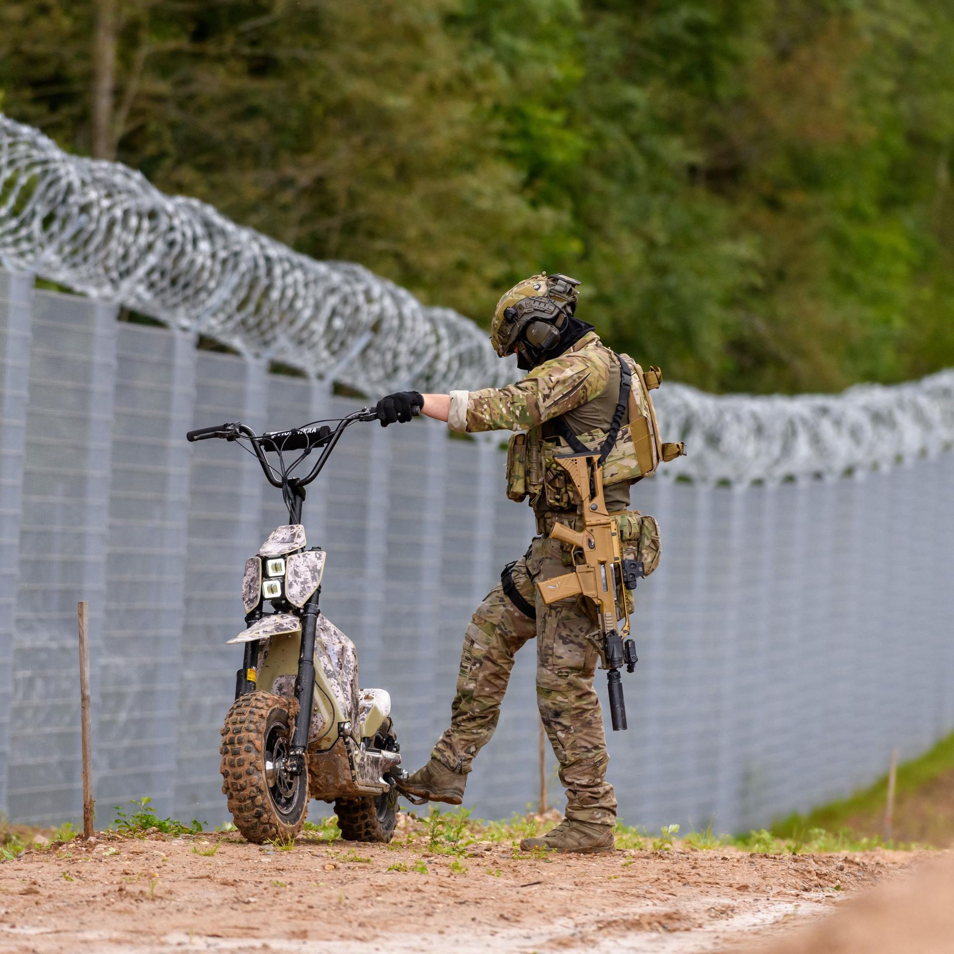 Ein Soldat der lettischen Grenzpolizei steht mit einem kleinen Motorrad am Zaun an der Grenze zu Belarus.