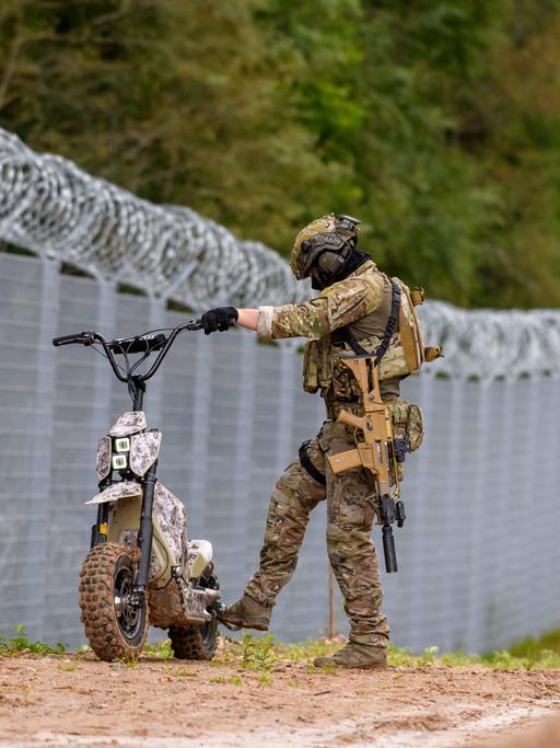 Ein Soldat der lettischen Grenzpolizei steht mit einem kleinen Motorrad am Zaun an der Grenze zu Belarus. Ein Soldat der lettischen Grenzpolizei steht mit einem kleinen Motorrad am Zaun an der Grenze zu Belarus.
