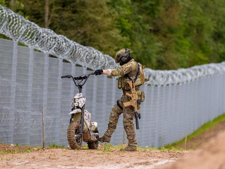 Ein Soldat der lettischen Grenzpolizei steht mit einem kleinen Motorrad am Zaun an der Grenze zu Belarus. Ein Soldat der lettischen Grenzpolizei steht mit einem kleinen Motorrad am Zaun an der Grenze zu Belarus.