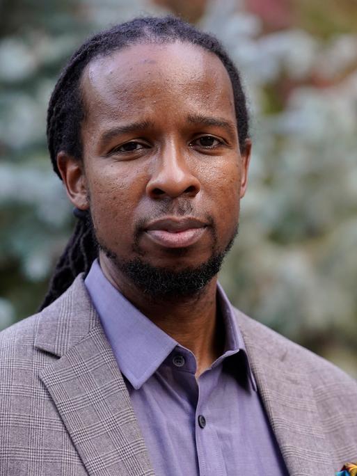 Porträtaufnahme des afro-amerikanischen Antirassimusforschers und -aktivisten Ibram X. Kendi.  Er blickt frontal in die Kamera, trägt einen grauen Anzug und die Haare lang zu einem Zopf zusammengebunden.