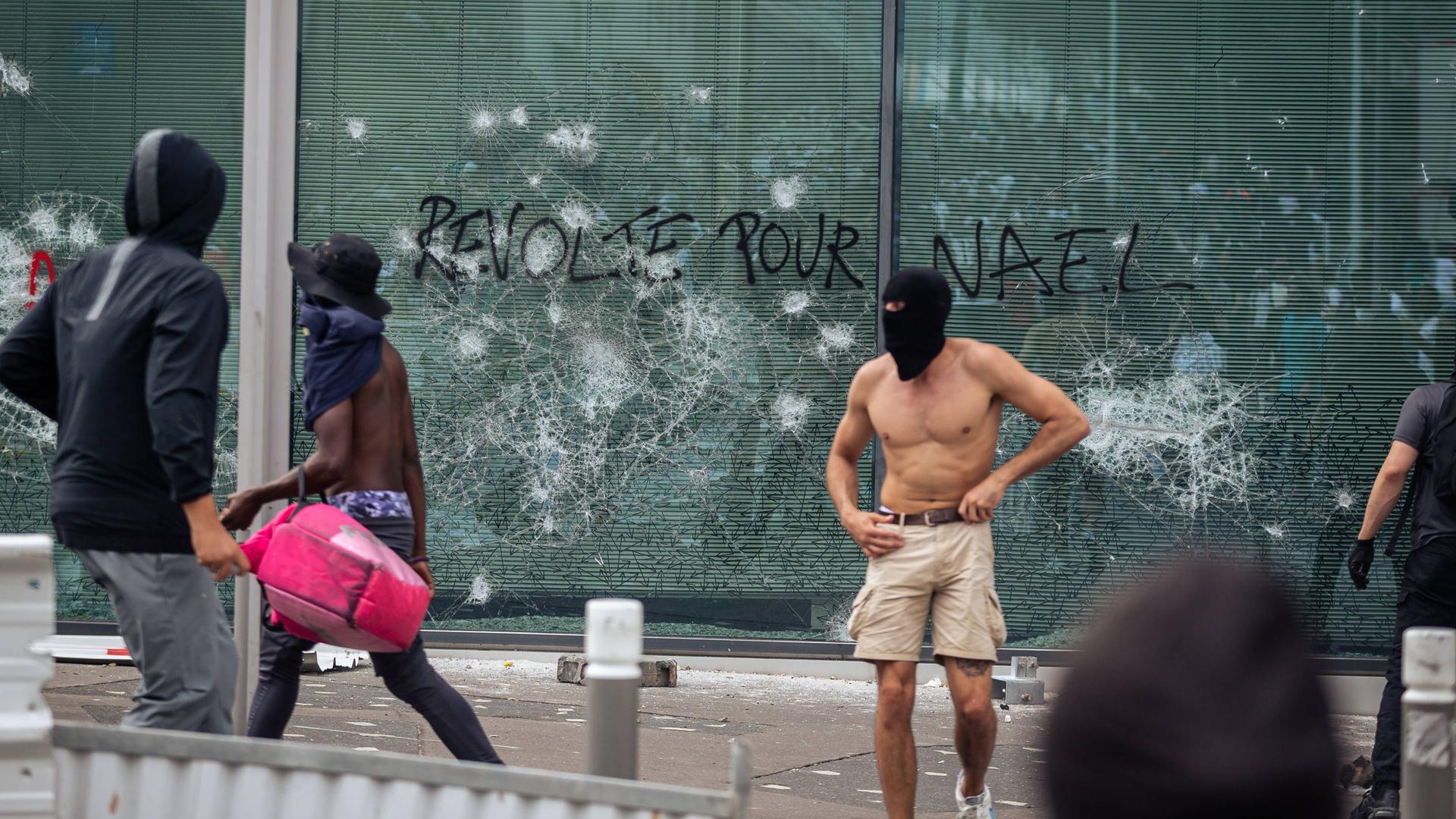 "Revolte für Nael" hat jemand auf die Scheibe eines Gebäudes in Paris geschrieben. Davor stehen mehrere junge Männer, die offenbar Teil der Auseinandersetzungen zwischen Polizei und Zivilisten sind, die gegen den gewaltsamen Tod des 17-jährigen Nahels durch einen Polizisten protestieren.