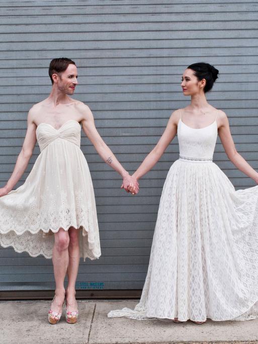 Ein männliches Gender-fluid-Model und eine junge Frau präsentieren Brautkleider vor einem Garagentor. Sie halten sich an der Hand und lächeln sich an.