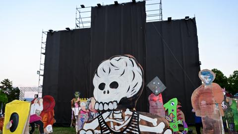 Von einem Metallgerüst hängen schwarze Stoffbahnen herunter. Auf der Wiese davor stehen Pappfiguren, die vorderste davon in Gestalt eines Skeletts mit Totenschädel. 