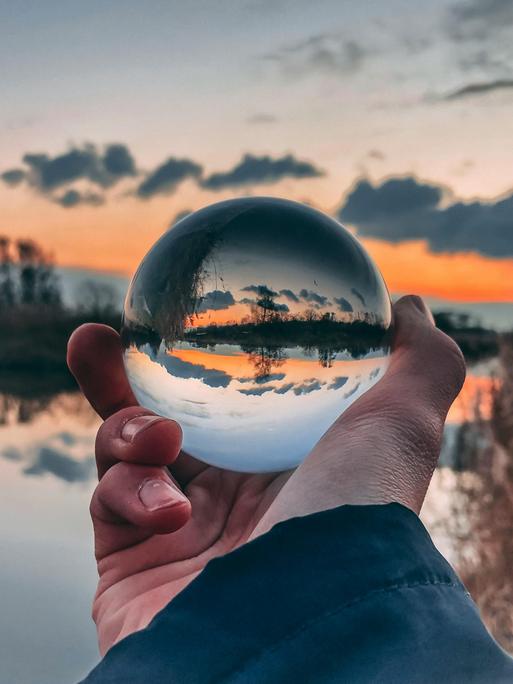 Ein Mann hält eine Glaskugel in der Hand, in der ein Sonnenuntergang mit umgekehrter Sicht der Landschaft erscheint.