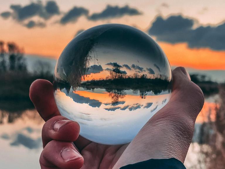 Ein Mann hält eine Glaskugel in der Hand, in der ein Sonnenuntergang mit umgekehrter Sicht der Landschaft erscheint.