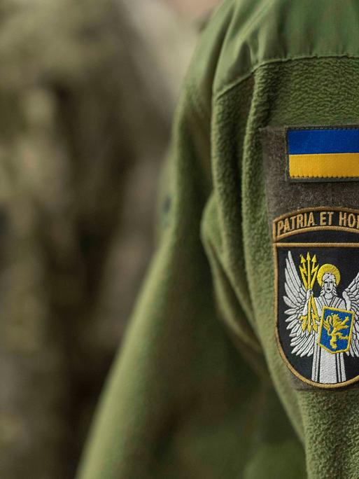 Zwei Personen tragen eine grünfarbene Uniform der ukrainischen Armee. Nur die Jacken sind zu sehen. Auf beiden ist je ein Aufnäher mit der ukrainischen Flagge genäht. Zwei Personen tragen eine grünfarbene Uniform der ukrainischen Armee. Nur die Jacken sind zu sehen. Auf beiden ist je ein Aufnäher mit der ukrainischen Flagge genäht.