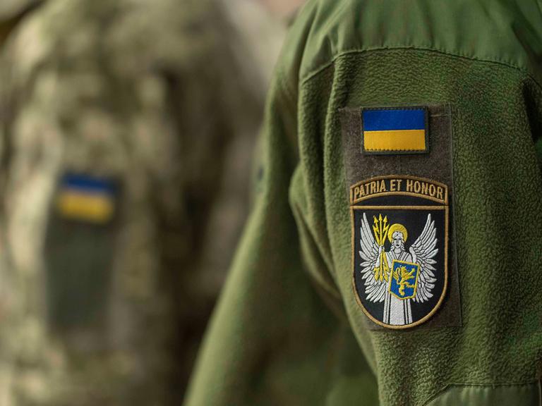 Zwei Personen tragen eine grünfarbene Uniform der ukrainischen Armee. Nur die Jacken sind zu sehen. Auf beiden ist je ein Aufnäher mit der ukrainischen Flagge genäht. Zwei Personen tragen eine grünfarbene Uniform der ukrainischen Armee. Nur die Jacken sind zu sehen. Auf beiden ist je ein Aufnäher mit der ukrainischen Flagge genäht.