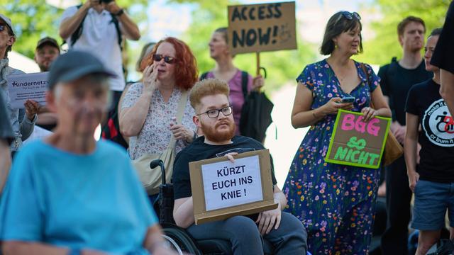 Mehrere Menschen stehen und sitzen nebeneinander. Sie halten Schilder zum Protest in die Höhe. Auf einem Schild steht "Kürzt euch ins Knie". Auf einem anderen Schild steht "Access Now". Das ist englisch und bedeutet "Zugang jetzt".