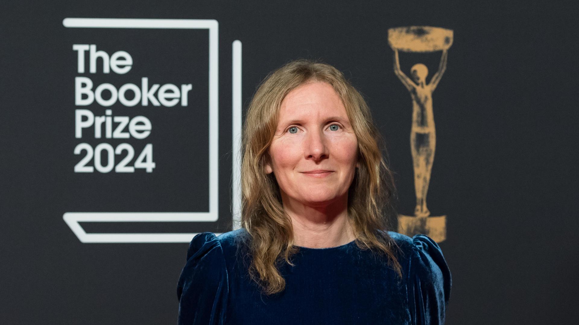 Samantha Harvey trägt ein dunkelblaues Samtkleid, steht vor der Fotowand des Booker Prize und lächelt in die Kamera