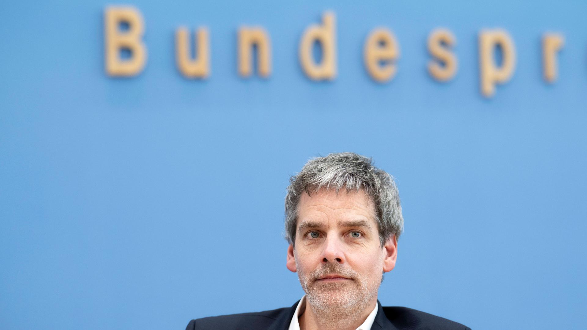 Bundespressesprecher Steffen Hebestreit sitzt vor einem blauen Hintergrund und schaut leicht an der Kamera vorbei. Im Hintergrund ist unscharf und angeschnitten das Logo der Bundespressekonferenz zu sehen.