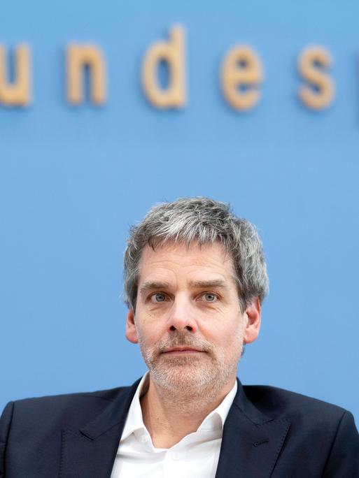 Regierungssprecher Steffen Hebestreit sitzt vor einem blauen Hintergrund und schaut leicht an der Kamera vorbei. Im Hintergrund ist unscharf und angeschnitten das Logo der Bundespressekonferenz zu sehen.
