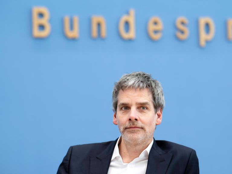 Regierungssprecher Steffen Hebestreit sitzt vor einem blauen Hintergrund und schaut leicht an der Kamera vorbei. Im Hintergrund ist unscharf und angeschnitten das Logo der Bundespressekonferenz zu sehen.