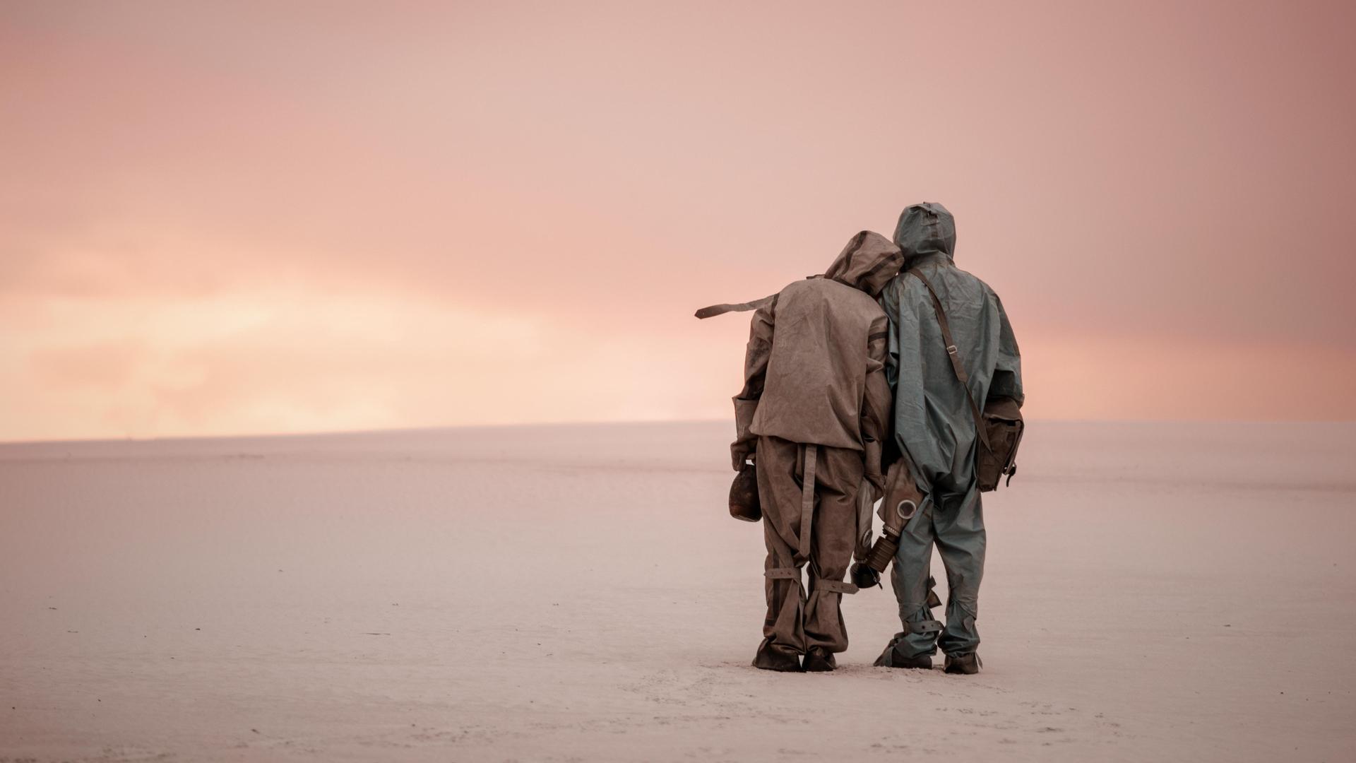 Zwei Personen in Schutzanzügen stehen zusammen in sandiger Landschaft, Gasmasken in den Händen.