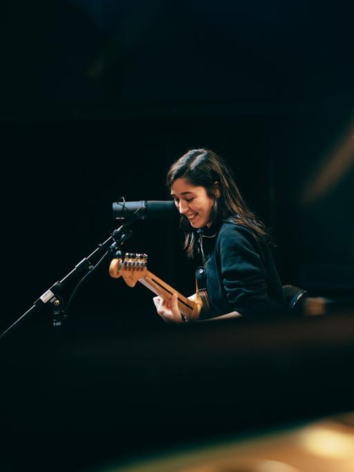 Eine junge Frau steht lachend vor einem Mikrofon mit ihrer Gitarre. Oska im Kammermusiksaal des Deutschlandfunks in Köln.