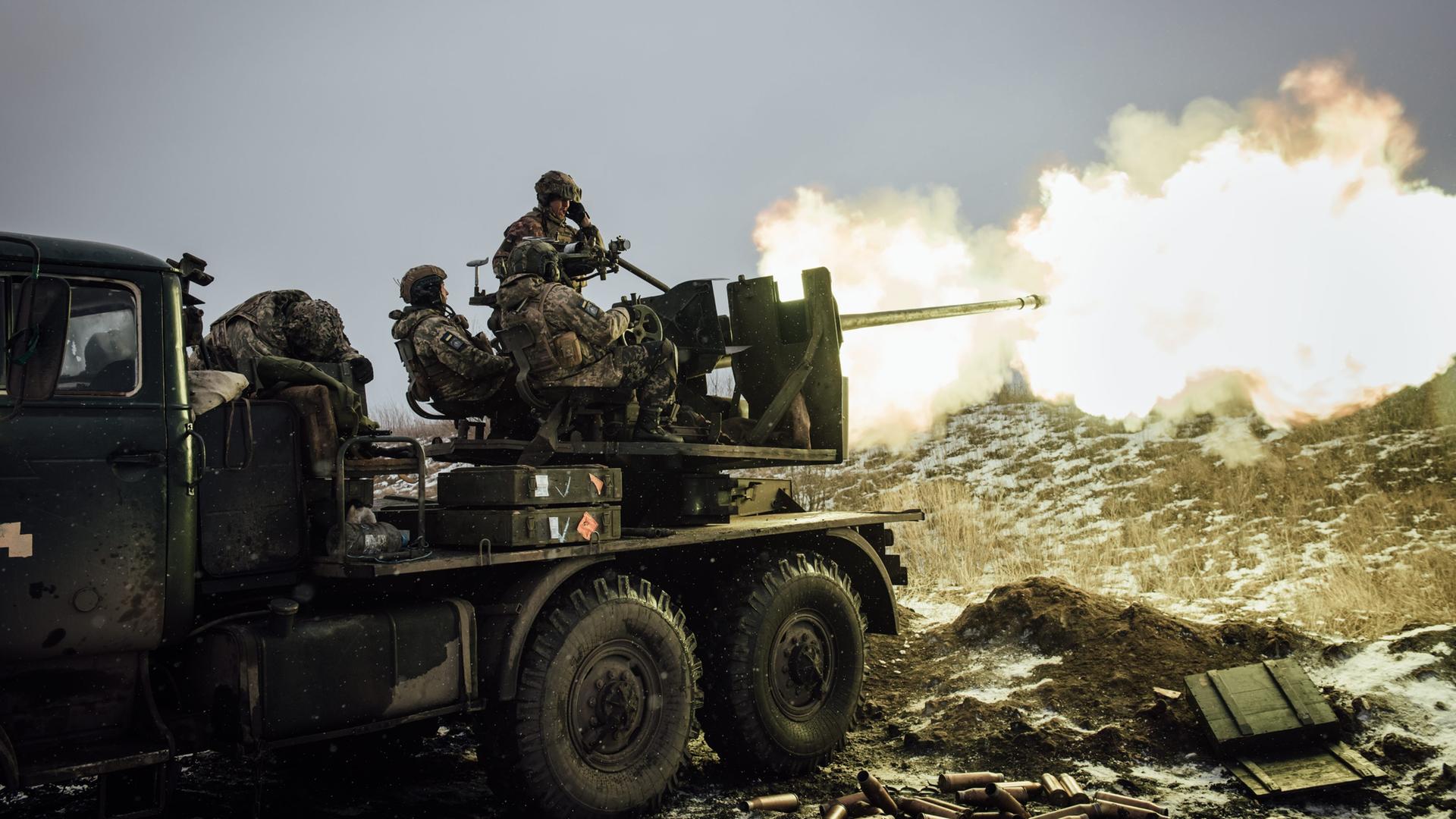 Ukrainische Soldaten sitzen auf einem Lastwagen mit Waffenvorrichtung. In der Ferne leuchtet Rauch.