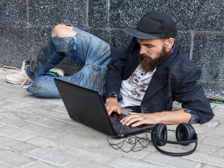 Ein junger Mann mit Baseballkappe liegt auf einem Bürgersteig auf dem Boden und bedient seinen Computer. 