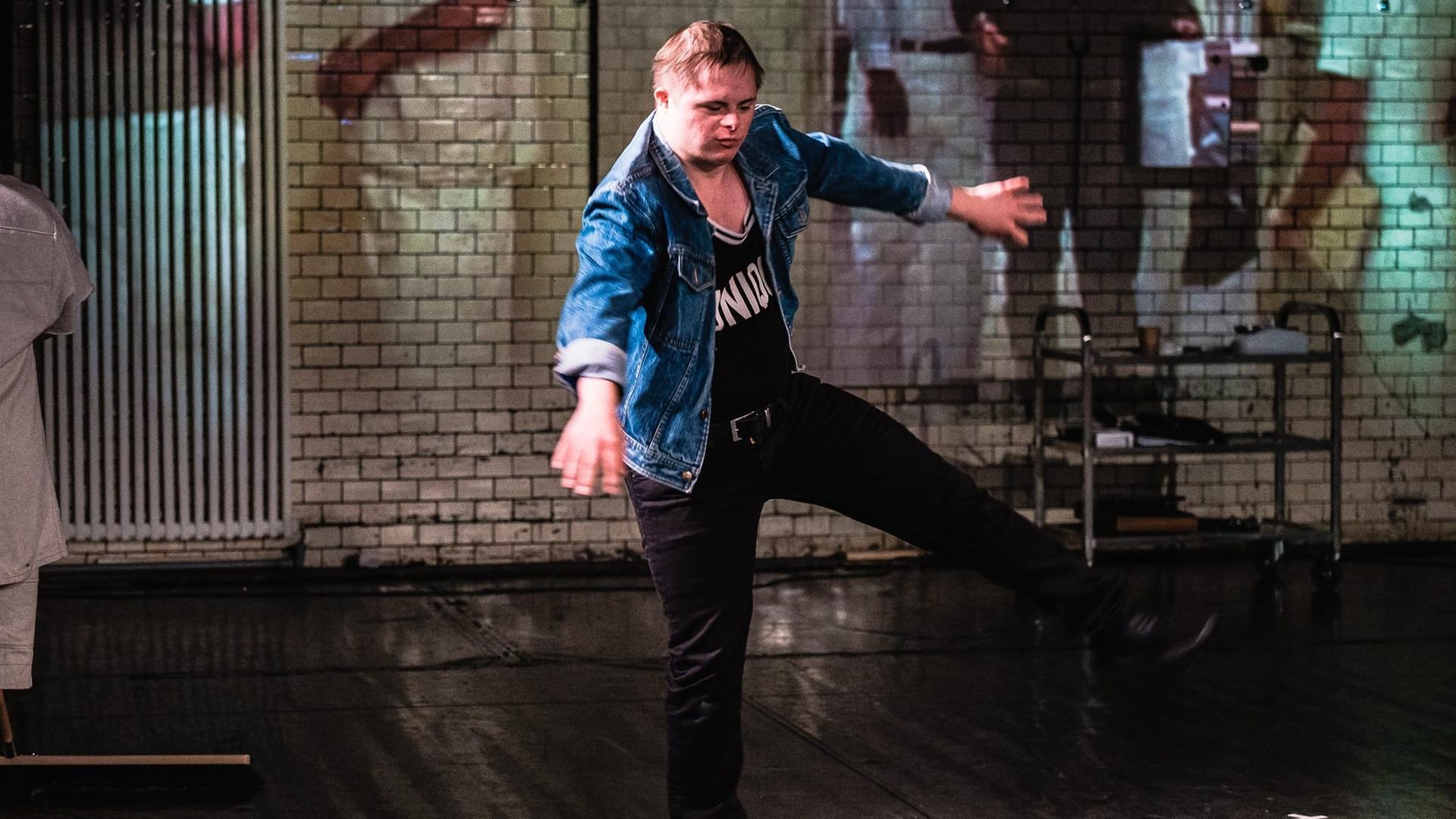 Ein Schauspieler in blauer Jeansjacke und dunklem T-Shirt balanciert auf einem Bein und rudert mit den Armen. Es ist ein Schauspieler der Theatergruppe RambaZamba in Berlin.
