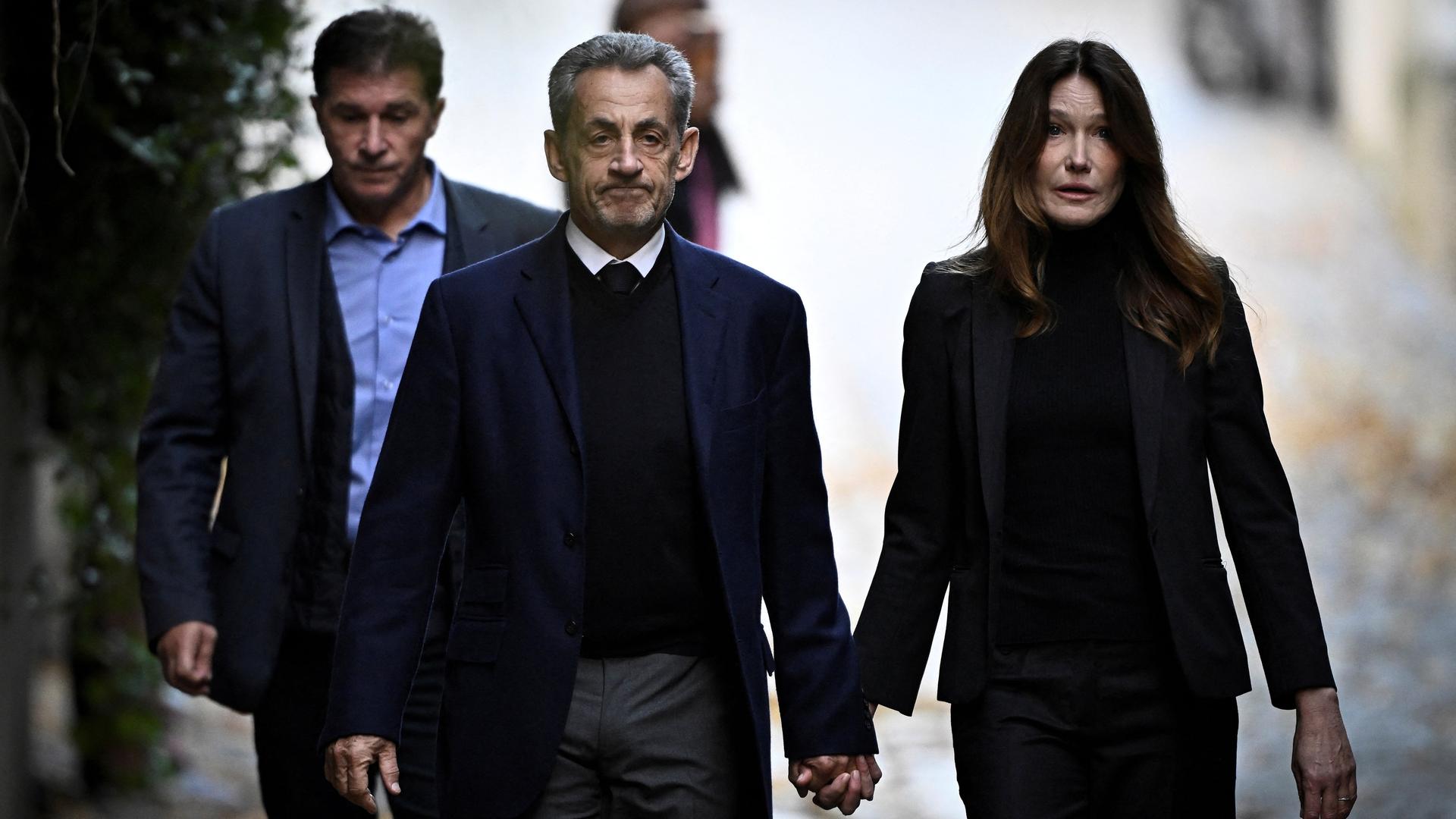 Sarkozy und seine Frau Carla gehen Hand in Hand und mit ernster Miene eine Straße entlang. Dahinter eine weitere Person. Man sieht sie von vorne. Sarkozy und seine Frau Carla gehen Hand in Hand und mit ernster Miene eine Straße entlang. Dahinter eine weitere Person. Man sieht sie von vorne.