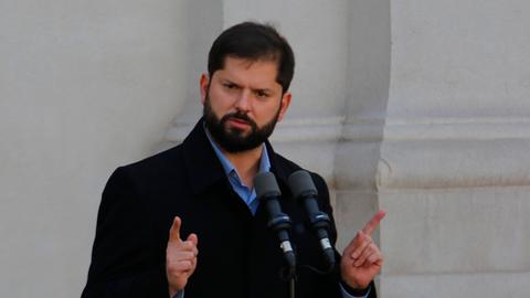 Ein Mann mit dunklem Vollbart steht im dunklen Mantel vor zwei Mikrofonen und hebt Hände samt Zeigefinger.