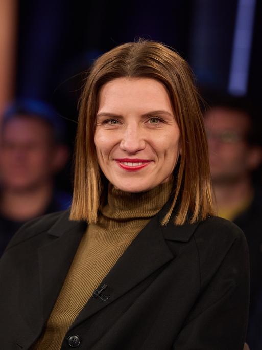 Magdalena Gössling, Autorin und ehemalige Ärztin, vor der Aufzeichnung der "NDR Talk Show" im Studio. Sie trägt einen braunen Rollkragenpullover und eine schwarze Jacke und lacht in die Kamera. Im Hintergrund ist das Publikum verschwommen auszumachen.