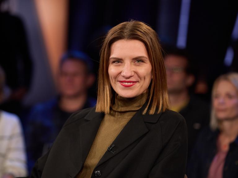 Magdalena Gössling, Autorin und ehemalige Ärztin, vor der Aufzeichnung der "NDR Talk Show" im Studio. Sie trägt einen braunen Rollkragenpullover und eine schwarze Jacke und lacht in die Kamera. Im Hintergrund ist das Publikum verschwommen auszumachen.