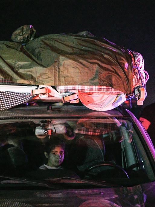 Flüchtlinge in einem Auto mit Gepäck auf dem Dach in der Nacht, ein Kind schaut heraus. Männer stehen neben dem Auto. 