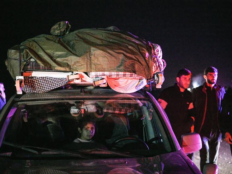 Flüchtlinge in einem Auto mit Gepäck auf dem Dach in der Nacht, ein Kind schaut heraus. Männer stehen neben dem Auto. 